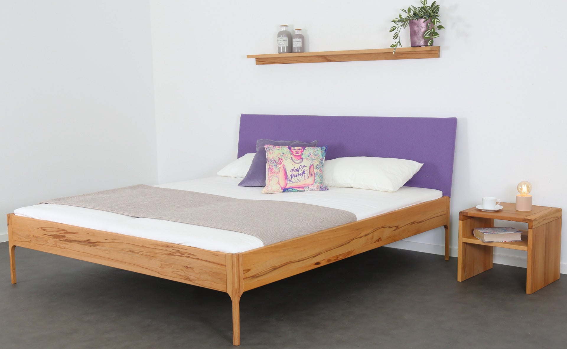 Schräge Frontalansicht von Bett Ona S in Kernbuche mit filigranen Füßen und rechteckiger Stofflehne in violett