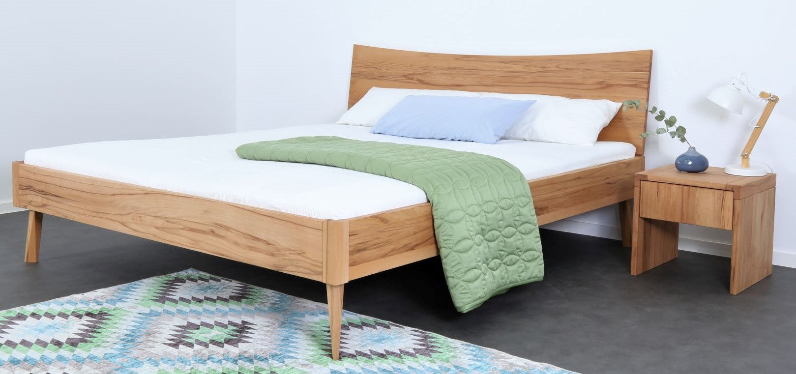 Schräge Frontansicht von Bett Tuxa 17 in Kernbuche mit konkaver Rückenlehne und filigranen Füßen